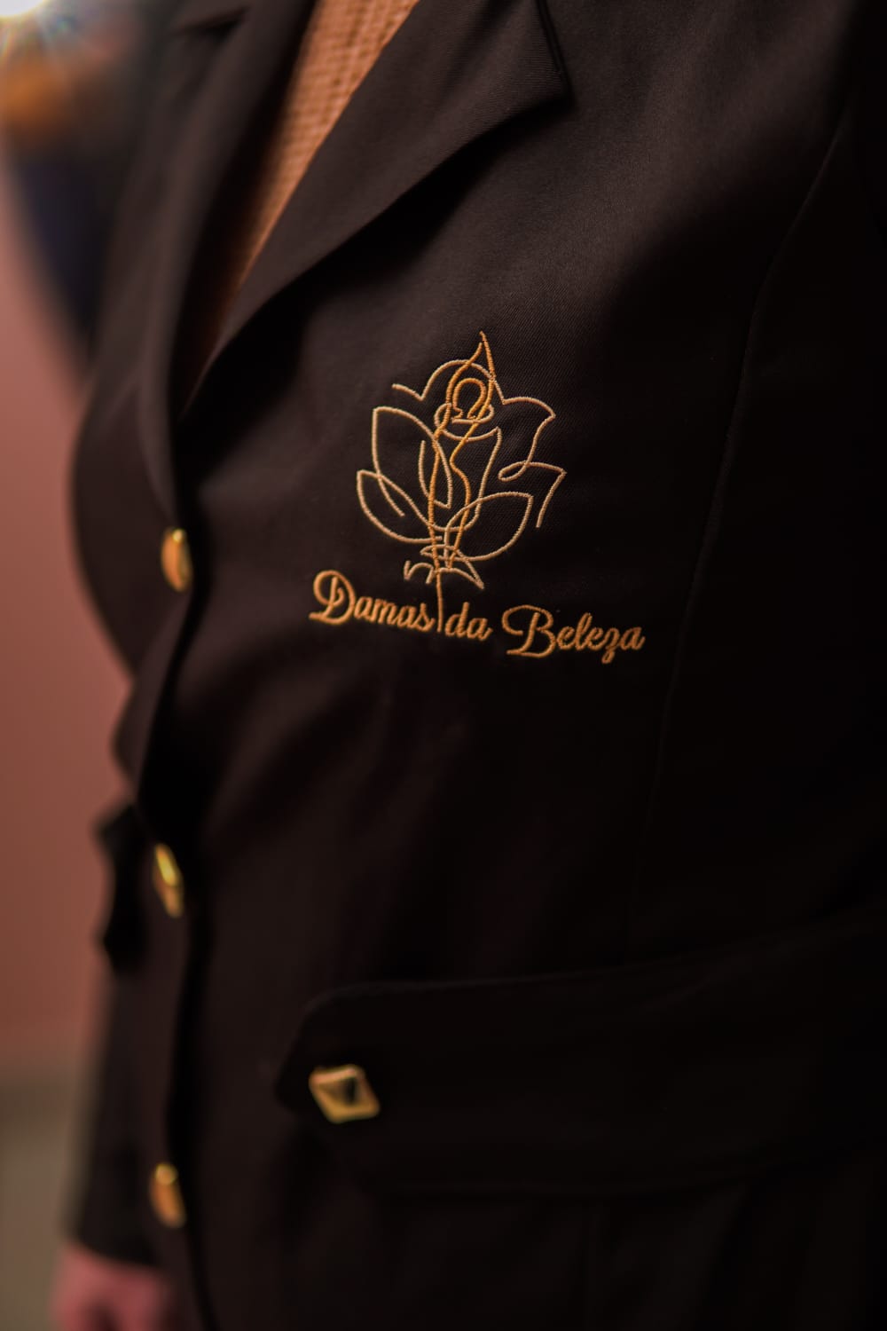Jaleco preto com o logo da Damas da Beleza bordado em fio dourado: uma magnólia com a silhueta de uma mulher no centro.